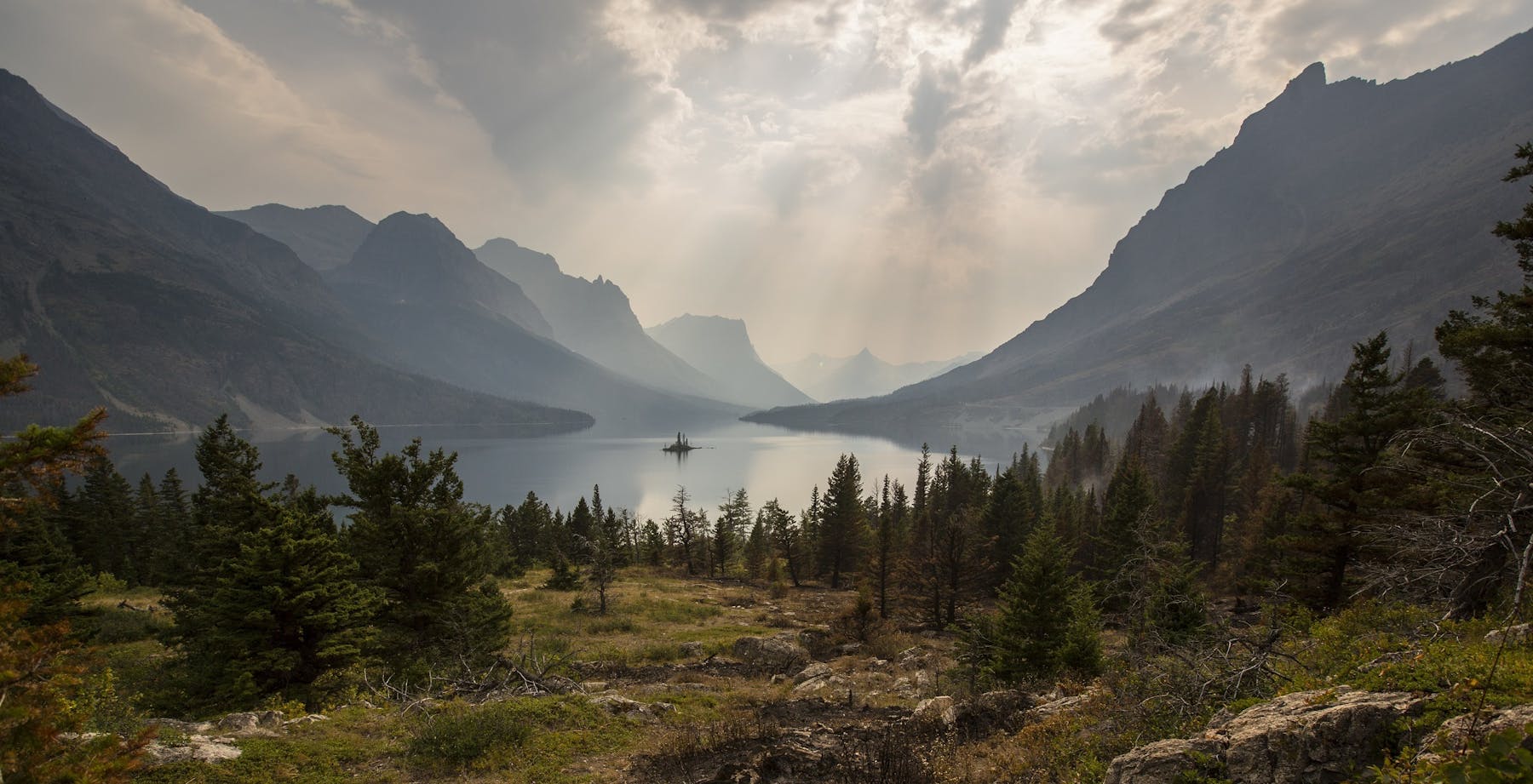 A still mountain lake ringed by evergreens and hazy peaks